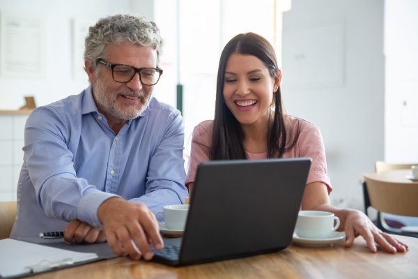Happy cheerful mature man and young woman sitting at open laptop, looking at display, watching content over cup of coffee and laughing. Medium shot. Digital communication concept